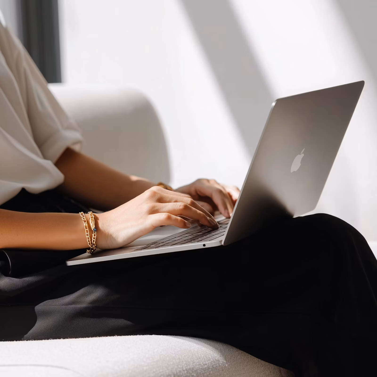 Person wearing a white shirt and black pants typing on a slim, silver Apple laptop while sitting on a light-colored couch with sunlight streaming in.